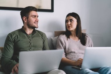 Ciddi genç çift, gündelik giysiler içinde dizüstü bilgisayarlarıyla yatakta oturuyor ve evde uzaktan kumandalı bir proje üzerinde çalışırken birbirlerine bakıyor.