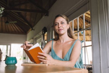 Düşük açılı, gülümseyen ve kameraya bakan genç bir kadın kahve fincanı defteri kalem ve günlükle gün boyu kafede oturuyor.