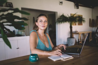 Evde çalışırken kameraya bakarken, açık dizüstü bilgisayarı ve gündelik kıyafetleriyle masada oturan serbest çalışan genç bir bayan.