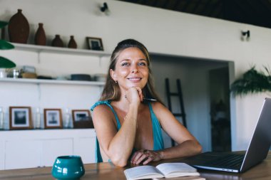Mavi bluzlu neşeli bayan serbest çalışan, gülümseyen ve kameraya bakan, dizüstü bilgisayarlı ve kahveli masaya oturan ve proje sırasında uzaktan notlar alan.