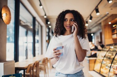 Kıvırcık saçlı, gülümseyen ve yüzünü çeviren pozitif Afro-Amerikalı kadın elinde kahve fincanı, cep telefonuyla gündüz vakti cam duvarlı, bulanık bir kafede konuşuyor.