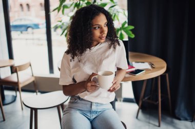 Düşünceli Afro-Amerikalı kadın dizüstü bilgisayarlı akıllı telefon defterli gözlüklü masada oturuyor ve elinde kahve bardakları, cam duvarları ve perdeleri olan bir kafede yeşil fabrikanın yanında duruyor.