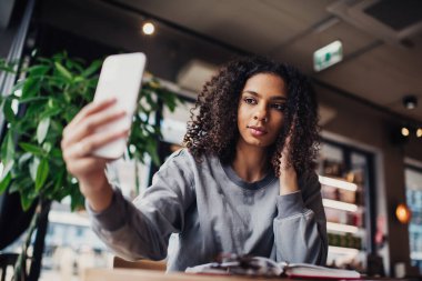 Gülümseyen genç bir kadının kıvırcık saçlarına elleriyle dokunması ve selfie çekerken cep telefonunun ekranına bakması ve bulanık kafedeki açık planlamacıyla masaya oturması.