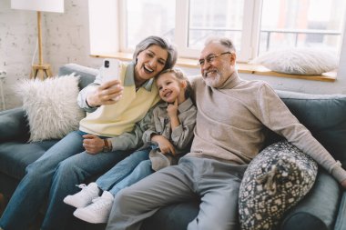 Aile içi giysiler içinde güzel sarışın bir kızla oturan ve evde vakit geçirirken kendi portresini çeken pozitif büyükanne ve büyükbaba.