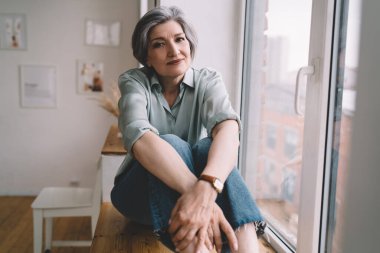 Gri saçlı, sıradan kıyafetler giyen, pencerenin kenarında oturan ve evde dinlenirken kameraya bakan son sınıf öğrencisinin huzurlu portresi.