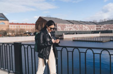 Güneş gözlüklü, sırt çantalı, cep telefonlu genç bir kadın yaz mevsiminde şehir binalarına karşı nehir kenarında demir korkuluklara bakarken görülüyor.