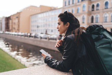 Siyah deri ceketli, sırt çantalı, cep telefonlu ve gün ışığında köprüde bulanık şehir binalarına yaslanmış genç turistlerin yan görüntüsü.