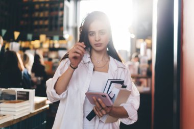 Üzerinde edebiyat ve akıllı telefon kitapları bulunan rafların yanında durup kameraya bakan rahat gömlekli genç bir etnik kadın.