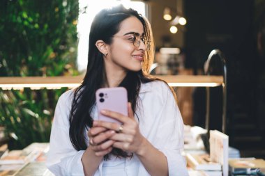 Güneş ışığına ve yeşil bitkilerle bulanık kitapçıya bakarken kapalı ellerde cep telefonu tutarak ve satılık yayınlarla masanın yanında duran gözlüklü pozitif genç bir kadın.