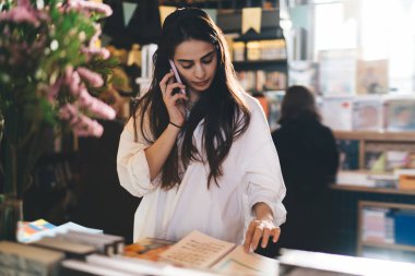 Kitaplığın yanında durup, modern kitapçıda okumak için ilginç bir kitap seçerken, akıllı telefondan konuşan, rahat giyinen genç bir kadın.