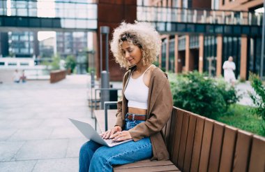 Tahta bankta otururken dizüstü bilgisayar kullanan ve internette proje üzerinde çalışan, günlük kıyafetli, kıvırcık saçlı Afro-Amerikalı genç bir kadının yan görüntüsü.