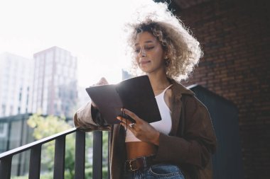 Merdivenlerde duran kıvırcık saçlı genç etnik kız öğrenci ve not defterinden notlar okurken tuğla duvarın yanındaki metal korkuluklara yaslanıyor.