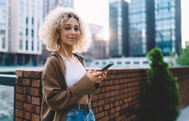 Palto ve kulaklıklı gülümseyen genç bir kız öğrenci. Tuğla duvara yaslanıyor ve cep telefonuyla mesaj yolluyor. Müzik dinlerken ve arka planda çok katlı binalar olan kameraya bakarken.