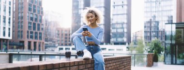 Afro-Amerikalı genç bir bayan gülümsüyor. Cep telefonunun ekranına bakıyor ve kaldırılmış bacağa yaslanıyor.