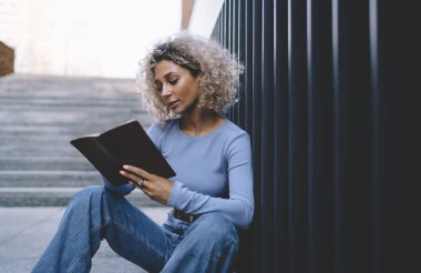 Okşanmış, kıvırcık saçlı, metal çitlerin yanında oturmuş sınav için bilgi okuyan Afro-Amerikan kız öğrencinin yan görüntüsü.