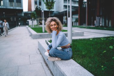 Genç siyahi kadın serbest kıyafetli, dizüstü bilgisayarlı, taş sınırında oturuyor ve modern şehre bakıyor.