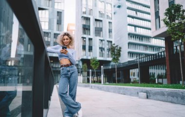 Kenarda duran ve cep telefonuna göz gezdiren genç bir kadın var. Ekrana bakıyor ve caddedeki cam parmaklıklara yaslanıyor.