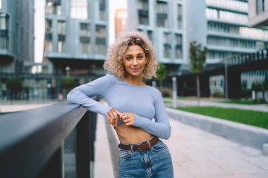 Gülümseyen kıvırcık saçlı kadın şehir merkezinde kaldırımda dikilip kameraya bakarken korkuluklara yaslanıyor.