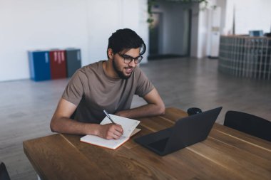 Gözlüklü, gülümseyen genç bir erkek kahve fincanı ile ahşap masada oturuyor ve internette bilgi ararken not defterine notlar yazıyor ve ofis kantinindeki dizüstü bilgisayara bakıyor.