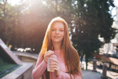 Çekici sarışın kadın taze limonata suyu içiyor ve şehir caddesinde güneş ışığında bulanık ağaçların yanında dururken kameraya bakıyor.