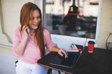 Gündelik giysiler içinde, dizüstü bilgisayarla masada oturan ve uzaktan çalışırken cep telefonuyla konuşan genç, pozitif bir bayan serbest yazar.