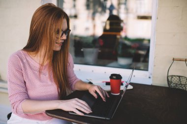 Kafedeki masada otururken dizüstü bilgisayarda yazan günlük giysiler ve gözlüklerle serbest çalışan genç bir kadın.
