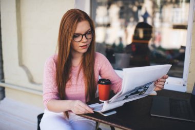 Konsantre olmuş kadın serbest çalışan elinde bir fincan kahve bilgisayarı akıllı telefonuyla masada otururken proje belgelerine bakıyor ve pencerenin yakınındaki kafedeki caddenin bulanık aynalı görüntülerini kontrol ediyor.