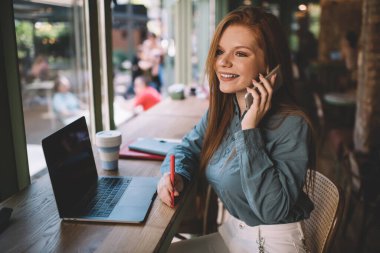 Kafe 'de oturmuş, akıllı telefondan konuşurken dizüstü bilgisayarla çalışan, şık kıyafetli, iyimser genç bayan girişimci.