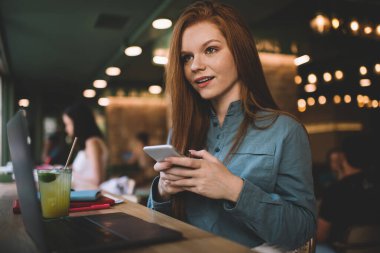 Gülümseyen ve yüzünü çeviren genç bir bayan elinde cep telefonu, elinde meyve suyu dolu bir dizüstü bilgisayarla masada oturuyor ve aydınlanmış kafede boş vakitlerin tadını çıkarıyor.