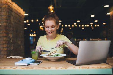Kafede dizüstü bilgisayarlı cam akıllı telefon günlüğüyle masada otururken aşağı bakan ve çatalla sağlıklı salata yiyen pozitif genç bir kadın.