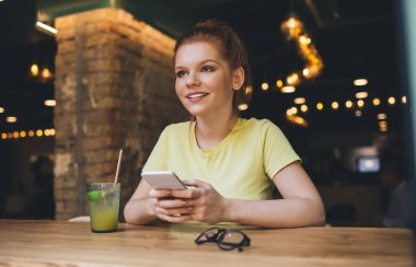 Ahşap masada bir bardak meyve suyu gözlüğü ile rahatlarken ve aydınlık kafede cep telefonu tutarken gülümseyen genç bir kadın gözlerini kaçırıyor.