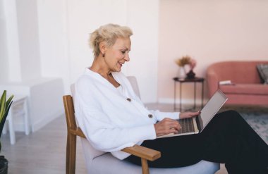 Orta yaşlı kadın serbest çalışanın yan görünüşü. Evde uzak bir proje üzerinde çalışırken çapraz bacaklı ve dizüstü bilgisayarla oturuyor.