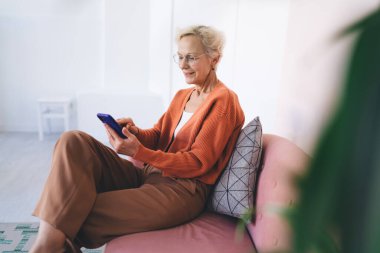 Oturma odasındaki kanepede yastığa yaslanırken, gözlüklü, cep telefonlu ve ekranda gezinen olgun bir kadının yan görüntüsü.