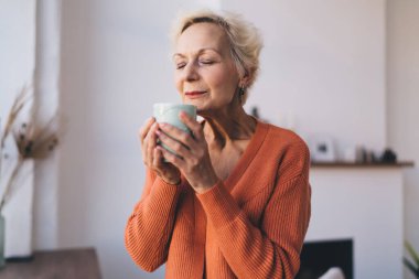 Sakin, sıcak süveter giyen, gözleri kapalı sıcak bir bardak tutan ve sıcak bir içecek içen, evdeki rahat oturma odasında eğlenen son sınıf öğrencisi bir kadın.