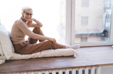 Ev kıyafetleri ve gözlüklü olumlu yaşlı bayan geniş pencereli yastıklarla yumuşak bir battaniyeye oturmuş kameraya bakıyor.