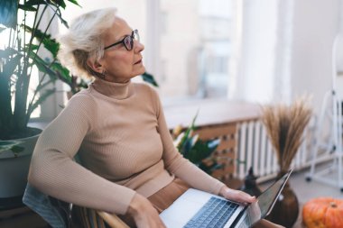 Kısa gri saçlı, sandalyede oturan ve dizüstü bilgisayarını kullanan düşünceli kadın serbest yazar. Hem de evdeki dinlenme odasında işe başlarken.