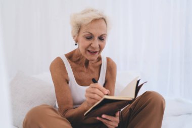 Gri saçlı, günlük kıyafetler giyen, yatakta oturan ve açık beyaz odadaki deftere bilgi yazan son sınıf öğrencisi.