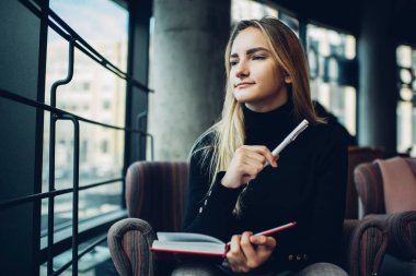 Kafe 'de oturmuş, pencerenin dışındaki şehir sokağına bakarken not defterine notlar yazan günlük kıyafetlerle konsantre olmuş genç kız öğrenci.