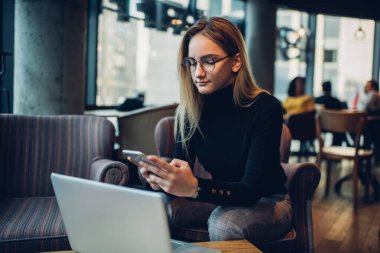 Modern kafeteryadaki koltukta oturmuş bilgisayarla uzaktan kumandalı cep telefonunu karıştıran, günlük kıyafetleri ve gözlükleriyle konsantre genç bayan uzaktan çalışan.