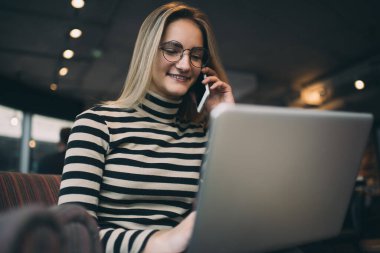 Çizgili balıkçı yaka kazaklı ve gözlüklü neşeli sarışın kadının düşük açılı görüntüsü kafede oturmuş bilgisayarda çalışırken telefonla konuşuyor.