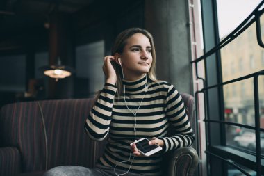 Düşük açılı, çizgili kazaklı, kulaklıklı, modern kahve dükkanında rahat bir koltukta oturan ve akıllı telefondan müzik dinleyen genç bir kadın.