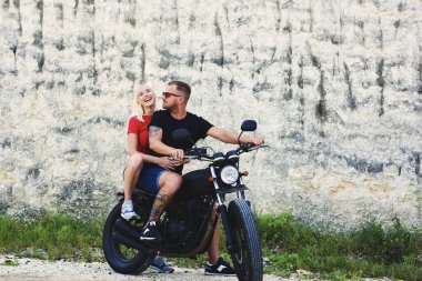 Havalı giyinmiş neşeli romantik çift yaz tatillerini motosiklet sürerek geçirmekten memnun, gülümseyen sarışın hippi kız erkek arkadaşıyla güneş gözlüklü maceranın keyfini çıkarıyor.