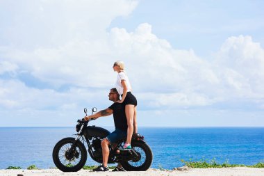 Çekici hippi kız motosikletli yakışıklı motosikletli erkek arkadaşıyla omuz omuza giderken okyanus kıyısında motosiklet süren sevimli bir çift birlikte macera yolculuğu yapıyor.