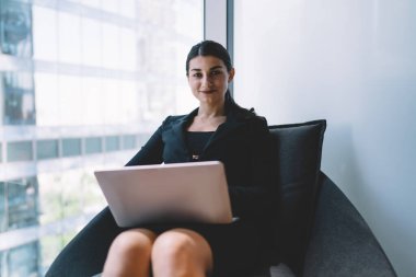 Resmi giysiler içinde gülümseyen genç bir kadın, pencerenin yanında dizüstü bilgisayarda çalışıyor, kanepeye oturuyor ve modern çalışma alanında kameraya bakıyor.