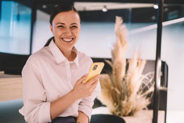 Neşeli bayan girişimci resmi giysiler içinde kameraya bakıyor ve gülümsüyor cep telefonunda internette geziniyor ve modern çalışma alanında çalışıyor.