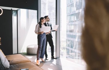 Cam duvarın yanında duran, şehir merkezine ve gökdelenlere bakan genç iş arkadaşlarının yan görüntüsü. Ofiste akıllı telefonlu koltukta oturan ekin işçisiyle birlikte çalışıyorlar.