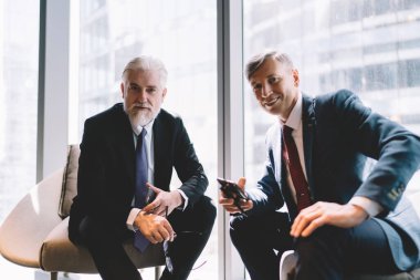 Orta yaşlı, takım elbiseli, gülümseyen ve kameraya bakan, modern ofisteki cep telefonlarıyla pencerenin yanında oturan üst düzey bir adam.