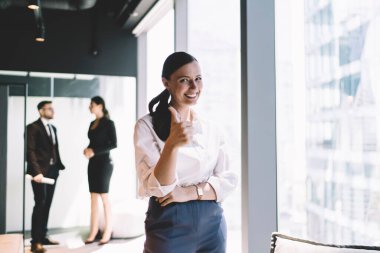 Resmi kıyafetli neşeli bayan yönetici. Gülümsüyor ve kameraya bakıyor. Çağdaş ofiste çalışırken başparmak hareketlerini gösteriyor.