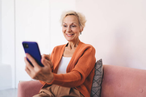 Positive aged female in orange cardigan smiling and looking at phone screen while resting on sofa and taking selfie during free time at home