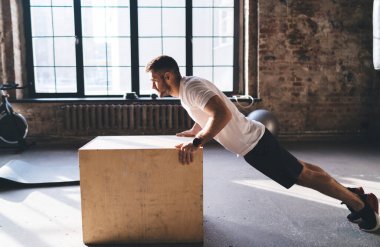 Spor giyim ve spor ayakkabıları giyen güçlü bir erkek sporcunun spor salonunda antrenman yaparken gözlerini kaçırarak tahta kutuda şınav çekerken görüntüsü.